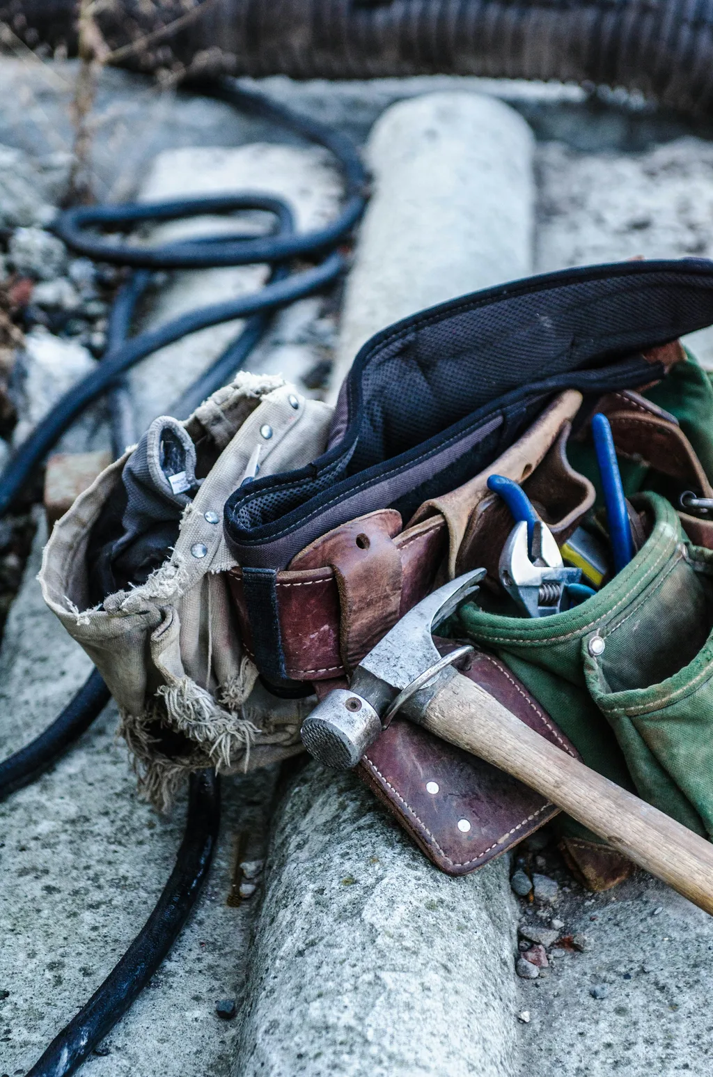 Worn leather tool belt holding a claw hammer and pliers resting on a concrete step