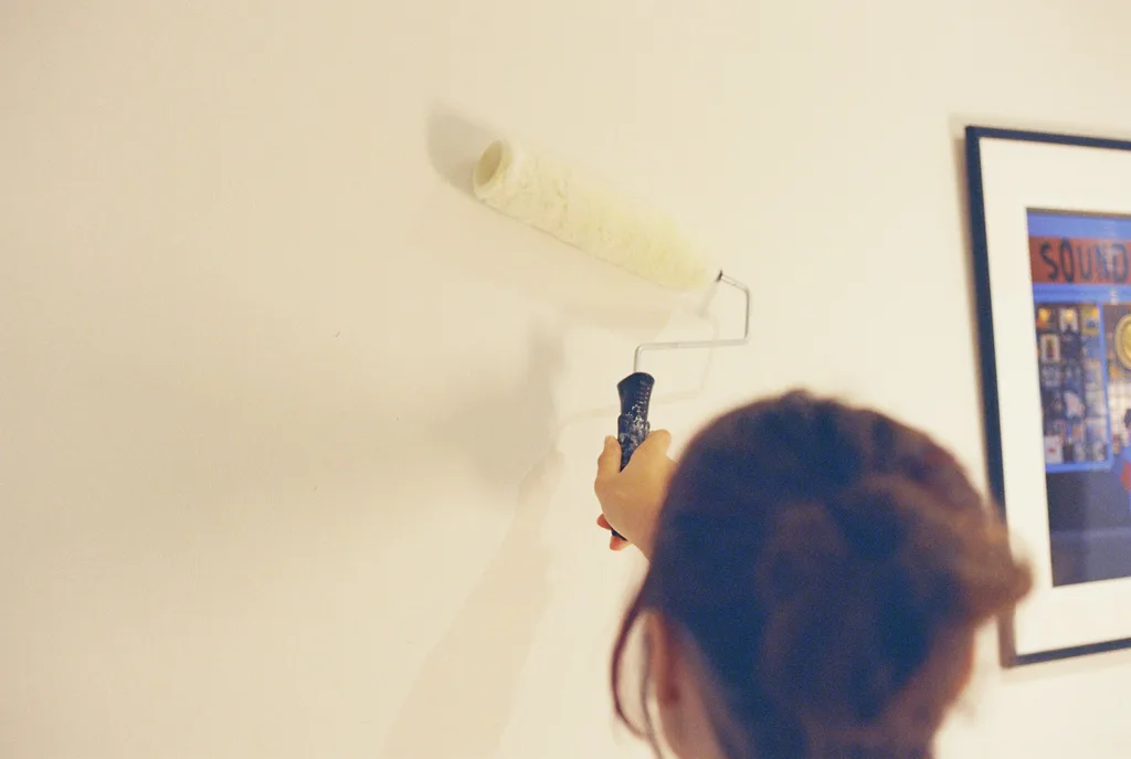 Person rolling fresh paint onto a pale interior wall beside a framed picture